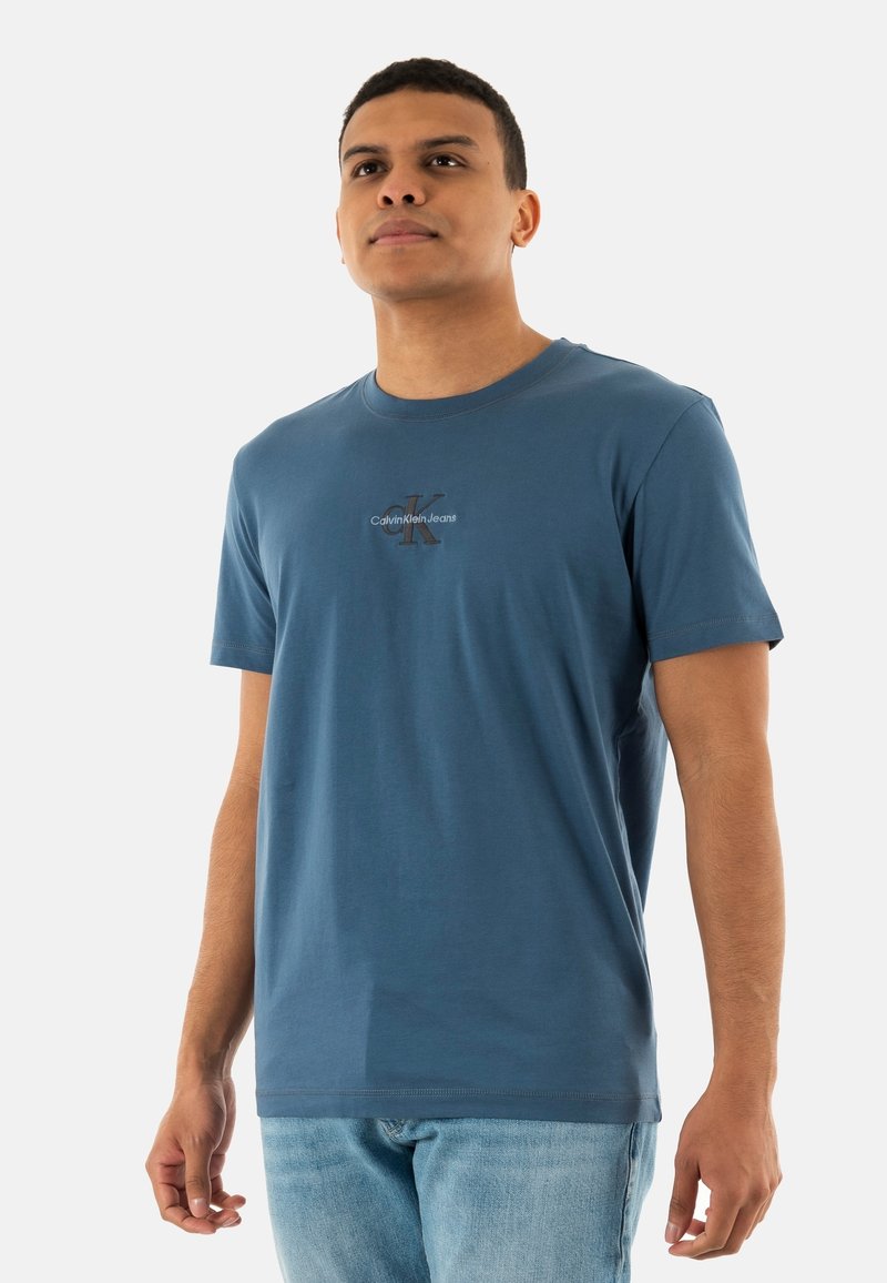 Camisa de algodón azul con mangas cortas, cuello redondo y un logo central. Textura suave, ajuste relajado. Combinada con jeans de un azul claro.