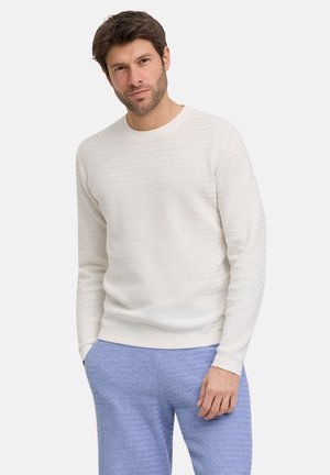 Mężczyzna z krótkimi brązowymi włosami i jasną bródką, ubrany w fakturowaną białą koszulę z długim rękawem oraz jasnoniebieskie fakturowane spodnie, stoi z jedną ręką w kieszeni.