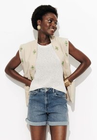 Gilet sans manches beige avec broderie verte, débardeur en tricot blanc et shorts en denim retroussés. Des boucles d'oreilles en or et des bracelets complètent le look.