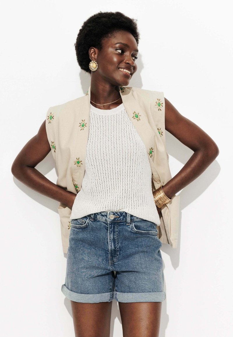 Gilet sans manches beige avec broderie verte, débardeur en tricot blanc et shorts en denim retroussés. Des boucles d'oreilles en or et des bracelets complètent le look.
