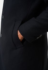 Black wool coat featuring a side pocket, button detailing on the cuff, and smooth texture. Close-up of sleeve and pocket area.