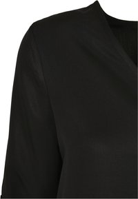 Blouse noire à col en V avec une texture lisse et mate. Présente un design épuré et un style minimaliste, mettant en valeur une silhouette modeste et ajustée.