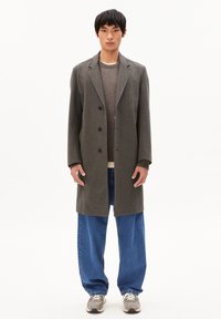Grauer, maßgeschneiderter Mantel mit Reverskragen und drei Knöpfen, kombiniert mit einem strukturierten braunen Pullover und lockeren blauen Jeans. Sneakers runden den Look ab.