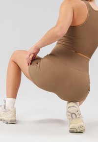 Ensemble court côtelé marron composé d'un crop top et d'une jupe assortie, avec une coupe ajustée. Le modèle porte des chaussettes blanches jusqu'à la cheville et des baskets beiges, dans une pose accroupie.