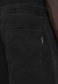 Svart bomullshose med en bakficka i rektangulär form och en vävd varumärkesetikett. Texturen verkar mjuk med subtila söm detaljer.