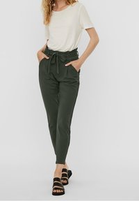 Pantalones verdes de talle alto con cinturón de tela, bolsillos laterales y pierna ajustada. Combinados con una camisa de manga corta en color crema y sandalias negras.