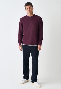 Bordeauxrotes Sweatshirt mit Rundhalsausschnitt und langen Ärmeln, kombiniert mit dunklen Jeans und weißen Sneakern. Glatte Textur, lässige Passform.