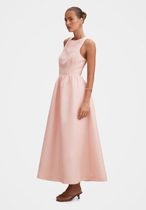 Donna che indossa un abito lungo fino al pavimento in raso rosa chiaro senza maniche, con collo alto e sandali con tacco basso, in piedi di profilo.