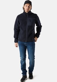 Giacca di fleece blu navy con zip anteriore, tessuto testurizzato, tasca sul petto e tasche laterali, abbinata a jeans blu e sneakers nere.