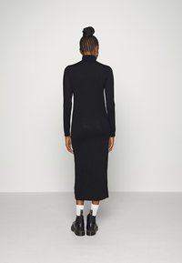 Schwarz, langärmeliges, figurbetontes Kleid mit hohem Kragen, aus geripptem Stoff, bis zur Mitte der Waden reichend. Trägt dazu schwarze, klobige Stiefel und weiße Socken.