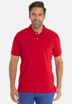 Homme portant un polo rouge et un pantalon bleu, souriant, se tenant devant un fond blanc uni.