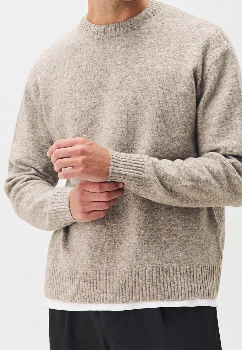 Homme ajustant la manche d'un pull en laine beige avec poignets et bas en côtes, portant un pantalon noir et une chemise blanche en dessous.
