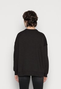 Persona con capelli corti scuri che indossa grandi orecchini a cerchio, una felpa nera larga e pantaloni neri, di fronte a un muro grigio chiaro semplice.