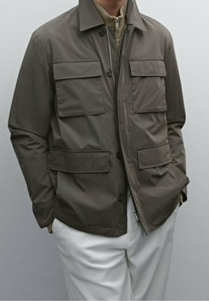 Man wearing olive green jacket with four flap pockets over beige zip-up sweater and white pants, hands in jacket pockets.