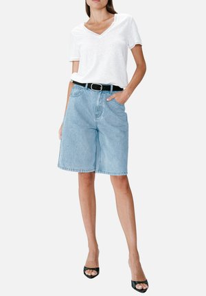 Femme portant un haut blanc à manches courtes avec col en V, un short en jean bleu clair jusqu'aux genoux avec une ceinture noire, et des talons noirs ouverts à l'avant, debout avec une main dans la poche.