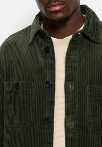Dark green corduroy jacket with a collared neck, front button closure, and two patch pockets. Features textured ribbed fabric and round black buttons.