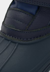 Botte bleu foncé avec une tige en cuir texturé, présentant des accents cousus et une semelle en caoutchouc flexible. Comprend des éléments de design superposés.