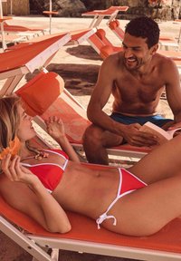 Bikini rouge avec accents blancs, présentant un haut triangulaire et un bas à nouer sur les côtés. Les accessoires incluent un collier avec des coquillages. Décor de plage.
