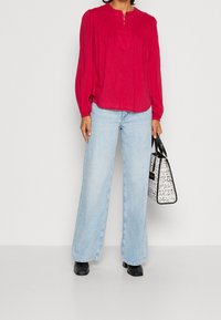 Röd långärmad topp med veck och knappdetaljer, kombinerad med ljusblå vida byxor och en mönstrad svart-vit handbag.