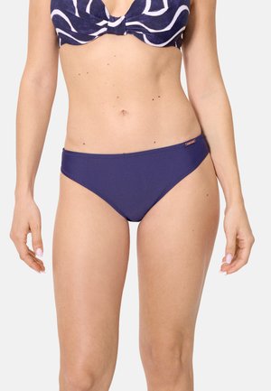 Mujer con braga de bikini azul marino y parte superior de bikini azul oscuro con estampado, de pie contra un fondo claro y liso.
