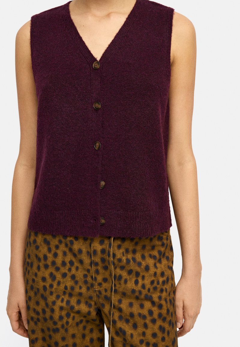 Burgundowy kardigan bez rękawów z dekoltem w kształcie V i pięcioma brązowymi guzikami z przodu. Miękka tekstura, połączony w plamkowanymi brązowymi spodniami.
