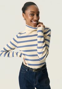 Mujer sonriente con el cabello recogido que lleva un suéter de cuello alto color crema con rayas azules y jeans oscuros, posando con la mano cerca de la barbilla.