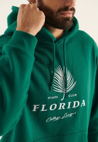 Sudadera verde de tela suave, con un gráfico de hoja de palma, el texto "FLORIDA" y capucha con cordón. Detalles de puños y dobladillo acanalados.