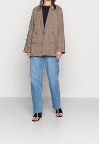 Osoba v hnědém kostkovaném oversized blejzru, černém topu, světle modrých širokých džínech a černých sandálech na podpatku s otevřenou špičkou stojící na šedé podlaze.