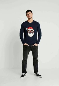 Marinblå stickad tröja med ett festligt tomteansikte med röd hatt och solglasögon. Matchas med mörka jeans och svarta sneakers.