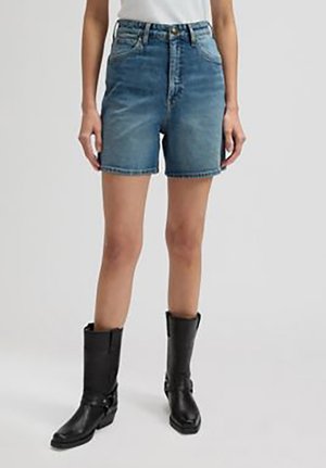 Kvinde iført blå cowboyshorts med høj talje og sorte støvler til midt på læggen, stående med hænderne afslappet ned langs siden.