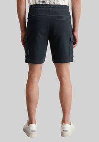 Shorts bleu foncé, arrivant au genou, en tissu texturé, avec une taille élastique et des poches cargo sur les côtés. Portés avec des baskets blanches. Vue de dos.