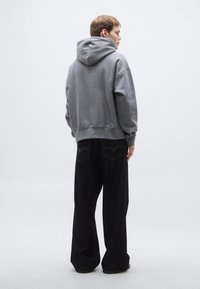 Szürke kapucnis pulóver laza szabással, fekete, széles szárú farmernadrággal párosítva, kontrasztos varrással és hátsó zseb részletekkel.