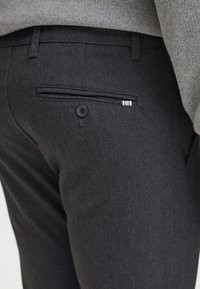 Temno sivo formalno hlače z teksturirano površino, ki imajo zadnji žep z gumbom in črtastim akcentom znotraj.
