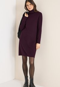 Vestido de punto morado con cuello alto, mangas largas y una silueta recta. Combinado con botas negras hasta el tobillo y medias negras transparentes.