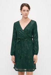 Robe en sequins verts avec décolleté en V, manches longues, taille cintrée et ourlet au niveau du genou. Le tissu a une apparence texturée et scintillante.