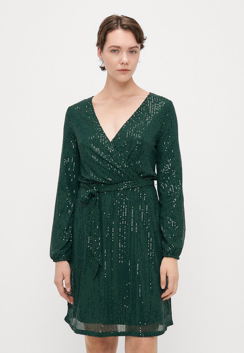 Robe en sequins verts avec décolleté en V, manches longues, taille cintrée et ourlet au niveau du genou. Le tissu a une apparence texturée et scintillante.
