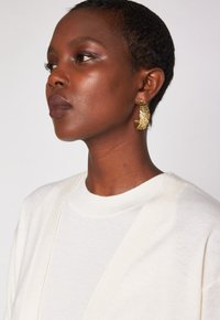 Boucle d'oreille en or texturé, dotée d'une surface martelée et d'un design ouvert. Portée avec un haut en tricot de couleur claire.