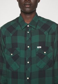 Zielona i czarna koszula z długim rękawem w kratę z kołnierzykiem zapinanym na guziki, dwiema kieszeniami na piersi oraz białą metką z logo na lewej kieszeni.