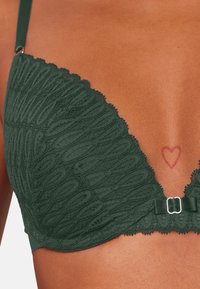 Czarny koronkowy stanik w ciemnozielonym kolorze z teksturowanym wzorem, falbaniastymi brzegami oraz kokardką z metalowym zapięciem z przodu. Widoczny tatuaż w kształcie serca.