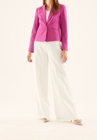 Fuksjowa marynarka z wyciętym kołnierzem i pojedynczym guzikiem, zestawiona z białymi szerokimi spodniami i beżowymi szpilkami z zaokrąglonym noskiem i ozdobami w formie kokardek.