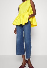 Mujer con top sin mangas amarillo con volantes, jeans cortos azul y zapatos de tacón alto de mimbre burdeos, de pie frente a un fondo liso.