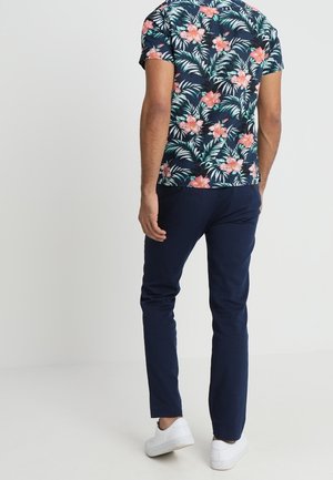 Mężczyzna noszący granatowe spodnie, białe sneakersy i ciemną, kwiecistą koszulę z krótkim rękawem stoi na jednolitym tle.