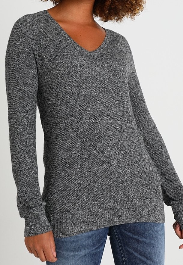 Femme vêtue d'un pull gris en tricot à manches longues et col en V, associée à un jean bleu, debout devant un fond clair uni.