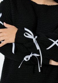 Schwarzer gestrickter Pullover mit weißen Schleifen aus Ripsband, die an den Ärmeln gebunden sind, die Arme verschränkt, um die Struktur und Designmerkmale zu zeigen.