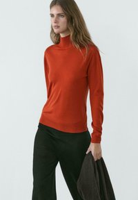 Frau mit langen braunen Haaren, die einen orangefarbenen Rollkragenpullover und schwarze Hose trägt, hält in ihrer rechten Hand ein zusammengefaltetes dunkles Kleidungsstück.