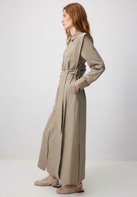 Beige, long-sleeved dress with a tied waist and side pockets. Fabric appears smooth, featuring button details along the front. Worn with patterned shoes.