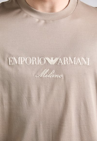 Bežinė medvilninė marškinė su išsiuvinėtą tekstu "EMPORIO ARMANI Milano" baltos spalvos, su mažuoju emblemu viršuje. Paprasta, lygi tekstūra.