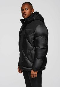 Giacca a vento nera con cappuccio, caratterizzata da un design trapuntato, tasche con zip e una toppa con logo sulla manica. Il materiale appare lucido e liscio.