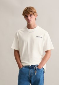 Witte katoenen T-shirt met korte mouwen en ronde hals, met "EARLY BIRDS" in donkere print op het linkerbovenborstgebied. Draagt blauwe spijkerbroek.