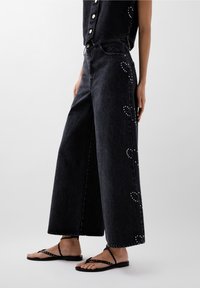 Zwarte denim jeans met wijde pijpen, versierd met hartvormige embellishments langs de buitenste naden, gecombineerd met zwarte platte sandalen.